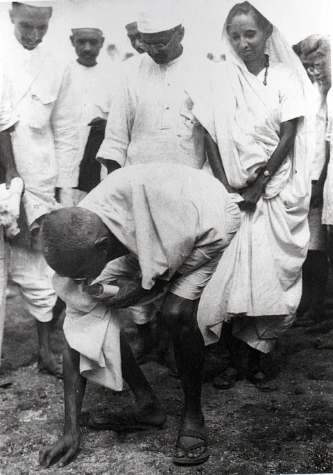 Gandhi, Tuz Yürüyüşü'nün sonunda Dandi sahilinde tuz toplarken; arkasında oğlu Manilal Gandhi ve Mithuben Petit görülüyor.